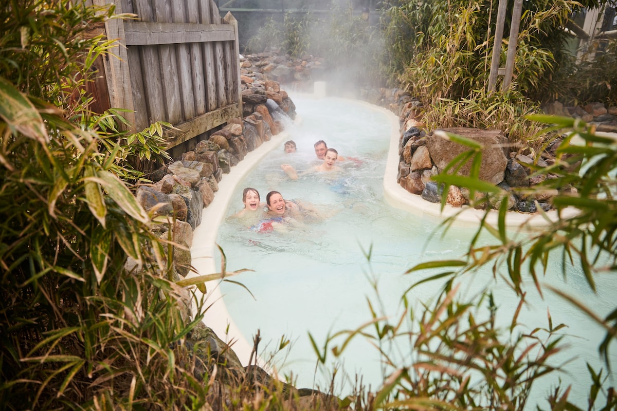 A serene hot spring is surrounded by smooth stones and lush bamboo. Steam rises gently from the water, creating a calming atmosphere. A winding path of clear water leads through the rocky landscape, inviting relaxation amidst nature.