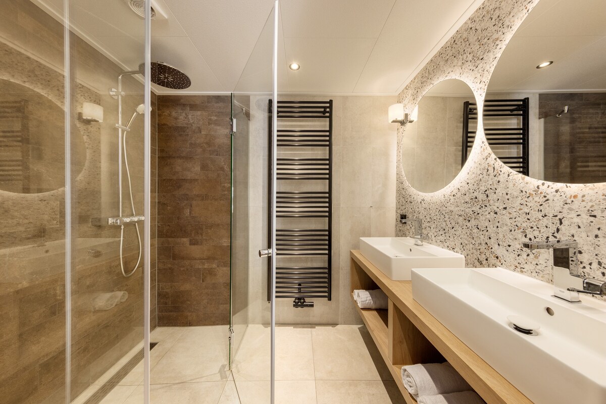 A spacious bathroom features dual vanities with modern sinks and elegant mirrors. A large walk-in shower with rainfall showerheads is visible alongside a towel radiator. Textured wall tiles provide a tasteful contrast against the light-colored flooring.