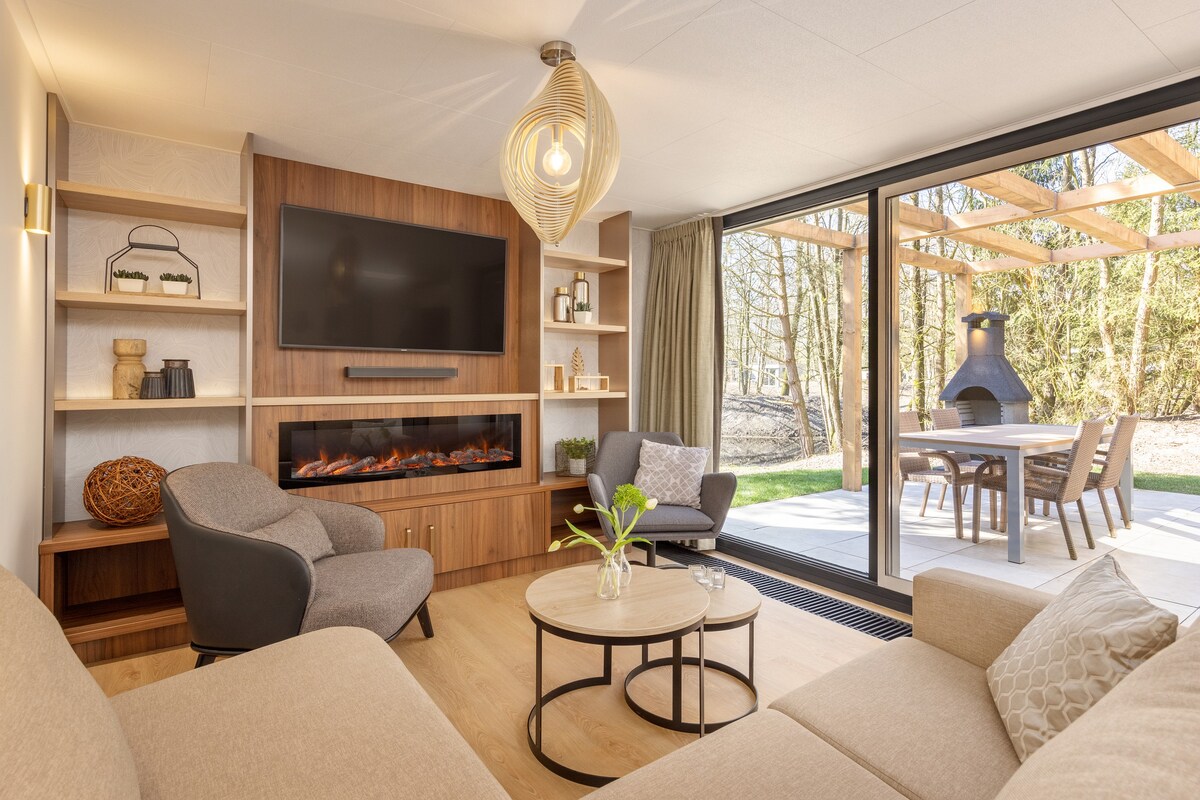 The living area features a cozy sofa and two armchairs, arranged around a circular coffee table. A flat-screen TV is mounted on the wooden wall unit, alongside decorative shelves. Natural light floods in through large glass doors, leading to an outdoor dining space under a covered terrace.
