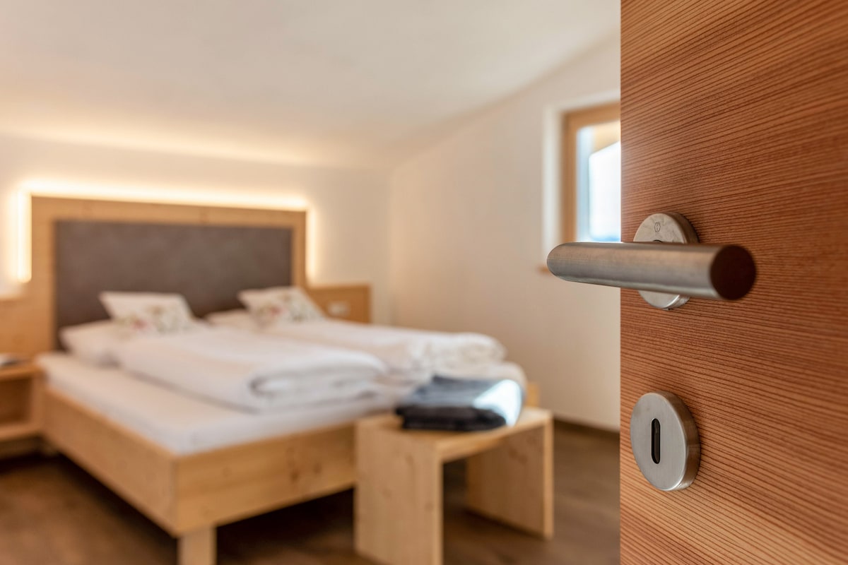 The entrance door provides a view into a bright bedroom featuring a spacious bed with white bedding and decorative pillows. Subtle lighting elements highlight the room's wooden accents, while a bedside table holds neatly folded items. Natural light filters through a nearby window.