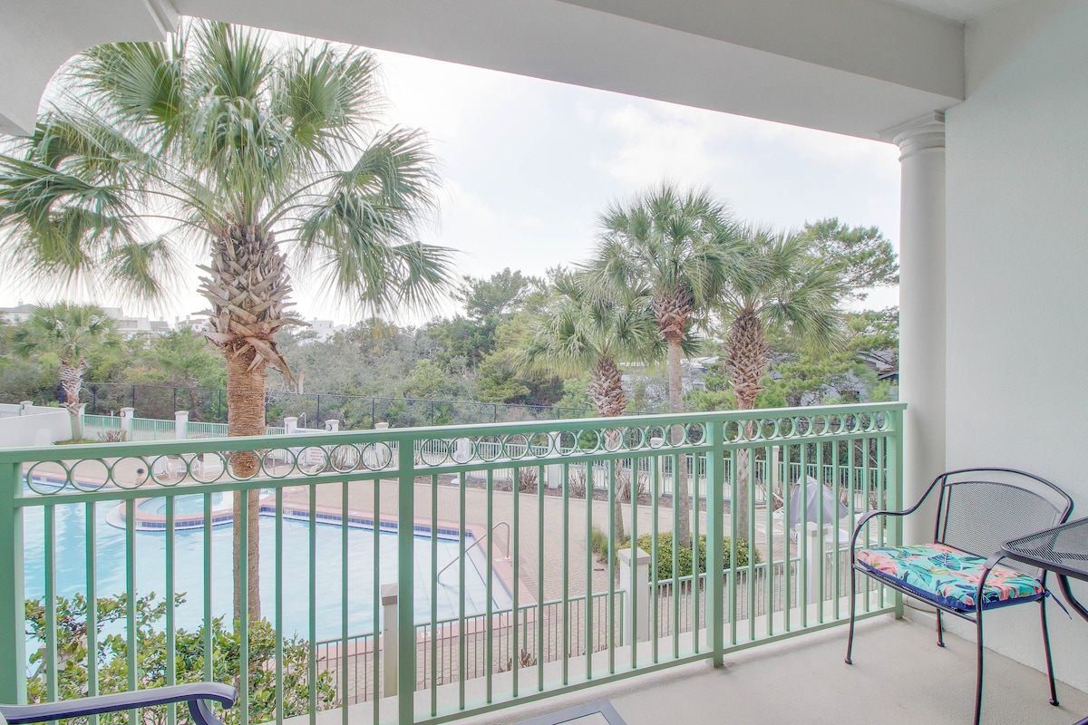 A private balcony showcases a view of swaying palm trees and a community pool below. Two black metal chairs with colorful cushions are positioned to enjoy the open air, while the surrounding greenery adds a relaxing touch to the scene.
