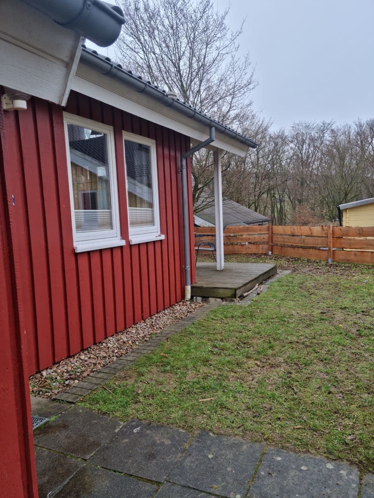 The exterior of the holiday home is shown, featuring red wooden siding and a small deck. A grassy area is visible alongside a stone pathway, and wooden fencing surrounds the yard, providing a sense of privacy. Trees in the background create a natural setting.