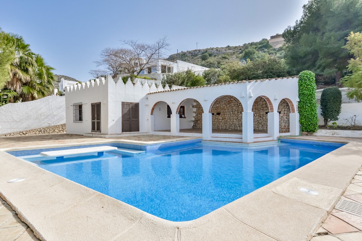 A serene swimming pool is surrounded by a well-maintained courtyard, featuring multiple arched structures that provide shade. Lush greenery borders the area, creating a tranquil atmosphere. The pool is clear, reflecting the blue sky and offers a combination of shallow and deep sections for relaxation and enjoyment.