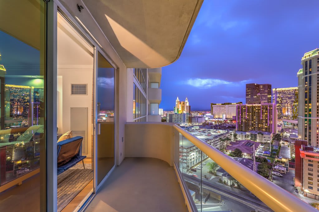 A private balcony offers sweeping views of the Las Vegas Strip, showcasing city lights against the evening sky. The space is bordered by glass sliding doors that connect indoors to the outdoor area, providing a seamless transition between the two.