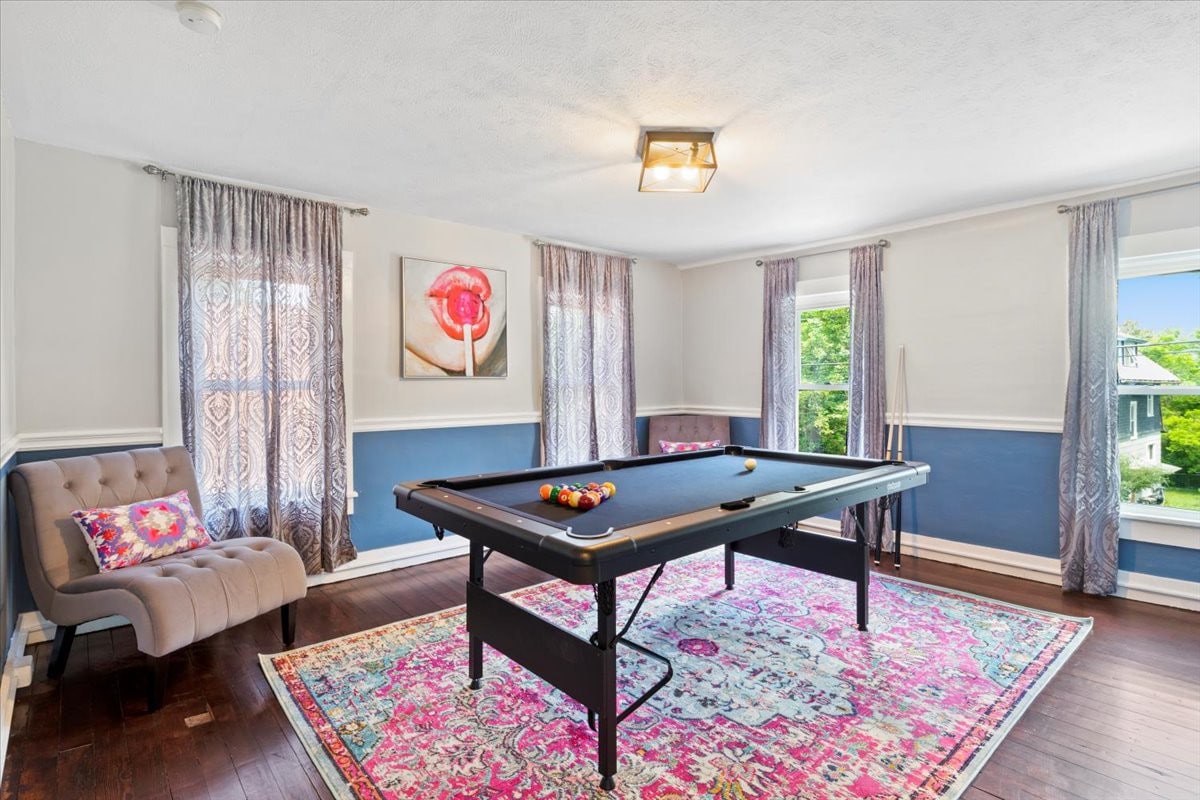 A well-lit room features a pool table centrally located on a colorful area rug. Large windows with patterned curtains provide natural light, while a cozy chair adds seating options. An artwork piece depicting a flower is displayed on the wall, enhancing the inviting decor.