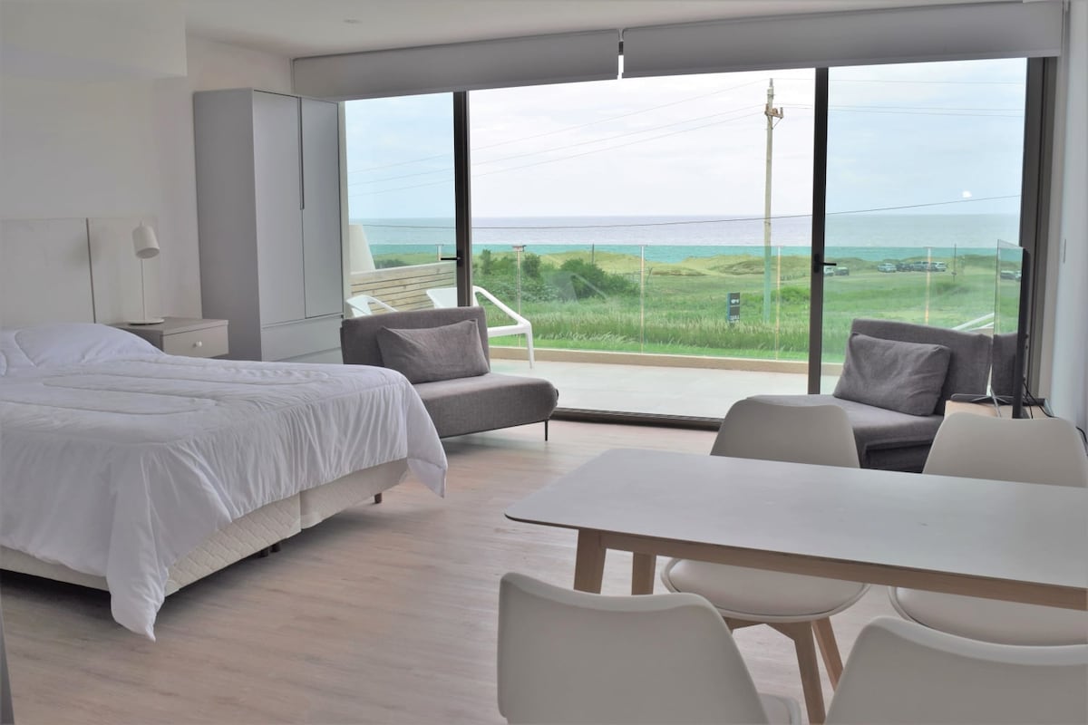 A spacious room features a large window offering a view of the ocean and surrounding greenery. A comfortable bed is set against one wall, while two armchairs and a dining table are positioned nearby. Natural light fills the space, creating a bright and inviting atmosphere.