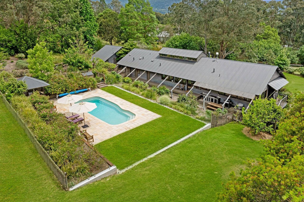 An aerial view showcases the spacious property featuring a salt-water pool, surrounded by well-maintained gardens and open green lawns. The house stretches along the back, with a large terrace offering shaded seating areas under an umbrella, creating a serene outdoor space.