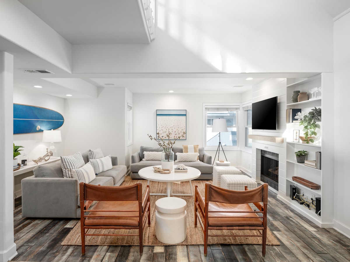 An open-concept living area features a spacious gray sectional sofa paired with light wooden chairs. A round coffee table at the center is surrounded by decorative accents, including a cozy area rug. Natural light floods through the windows, highlighting the modern decor and artwork.