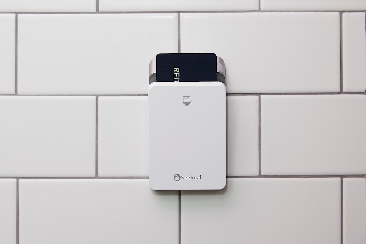 A sleek white card reader is mounted on a tiled wall with a variety of white tiles arranged in a grid pattern. The reader holds a black card, positioned at the top, indicating its use for access control.