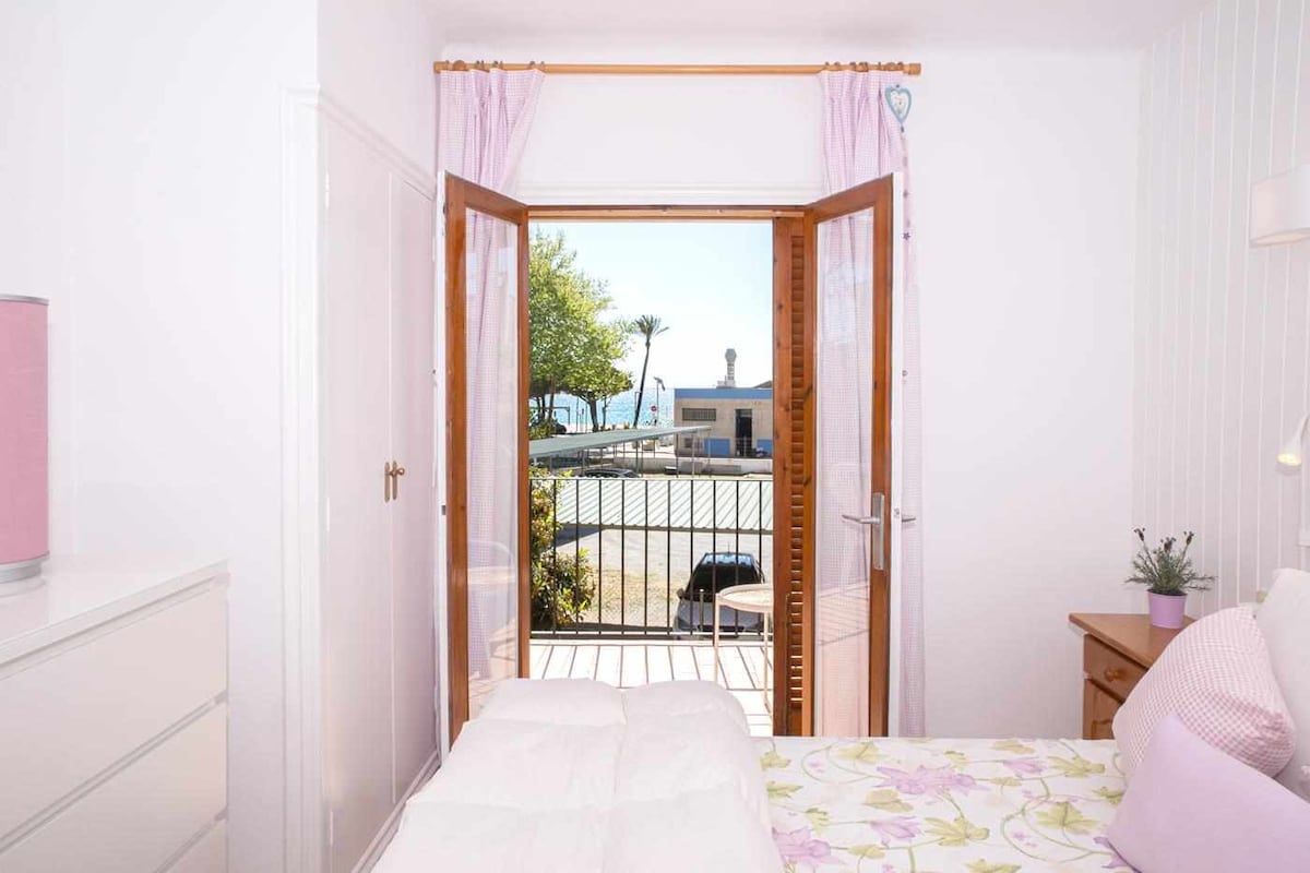 A bedroom features double doors opening to a terrace with sea views. Soft sunlight fills the space, illuminating the white walls and floral bedding. A dresser and bedside table complement the cozy atmosphere, while curtains gently frame the doorway, creating a serene setting.