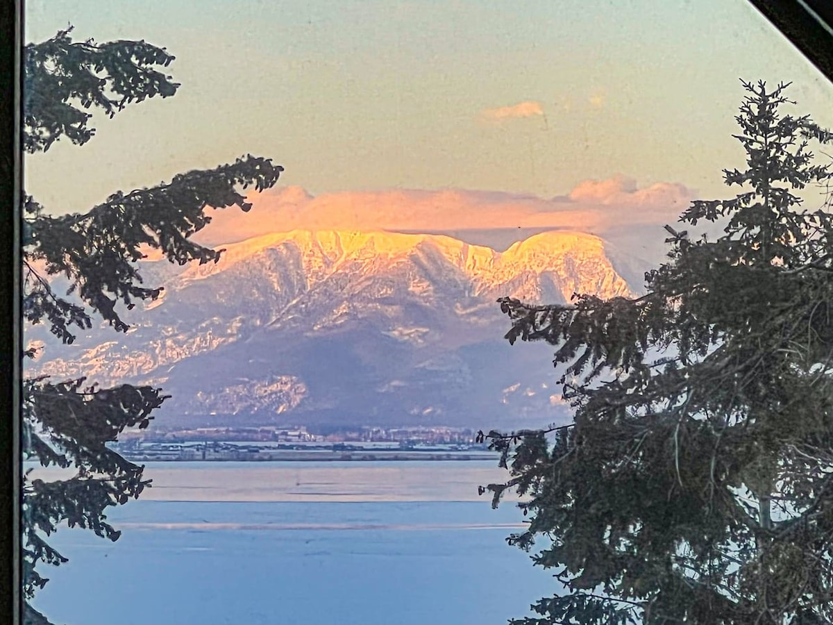 A scenic view of snow-capped mountains is framed by evergreen trees. The peaks are bathed in soft sunlight, contrasting against the serene lake below, which reflects the clear sky. This tranquil landscape highlights the natural beauty surrounding the property.