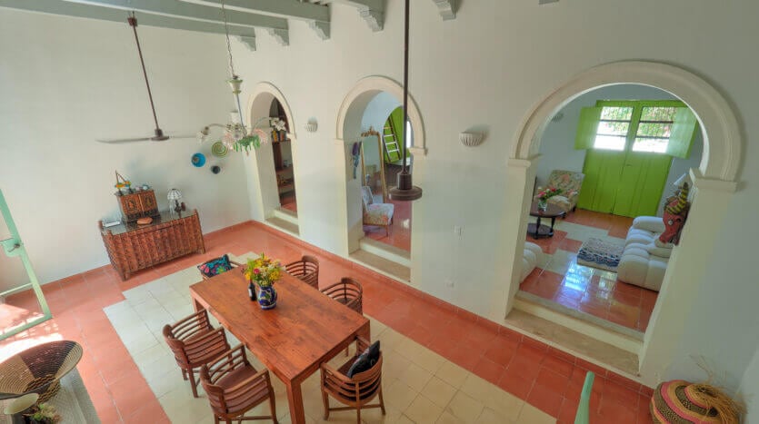 The airy living space features a large wooden dining table surrounded by multiple chairs. Arched doorways lead to comfortable seating areas, with vibrant decor visible in the background. A ceiling fan and decorative light fixtures enhance the room's ambiance, complemented by white walls and colorful accents.