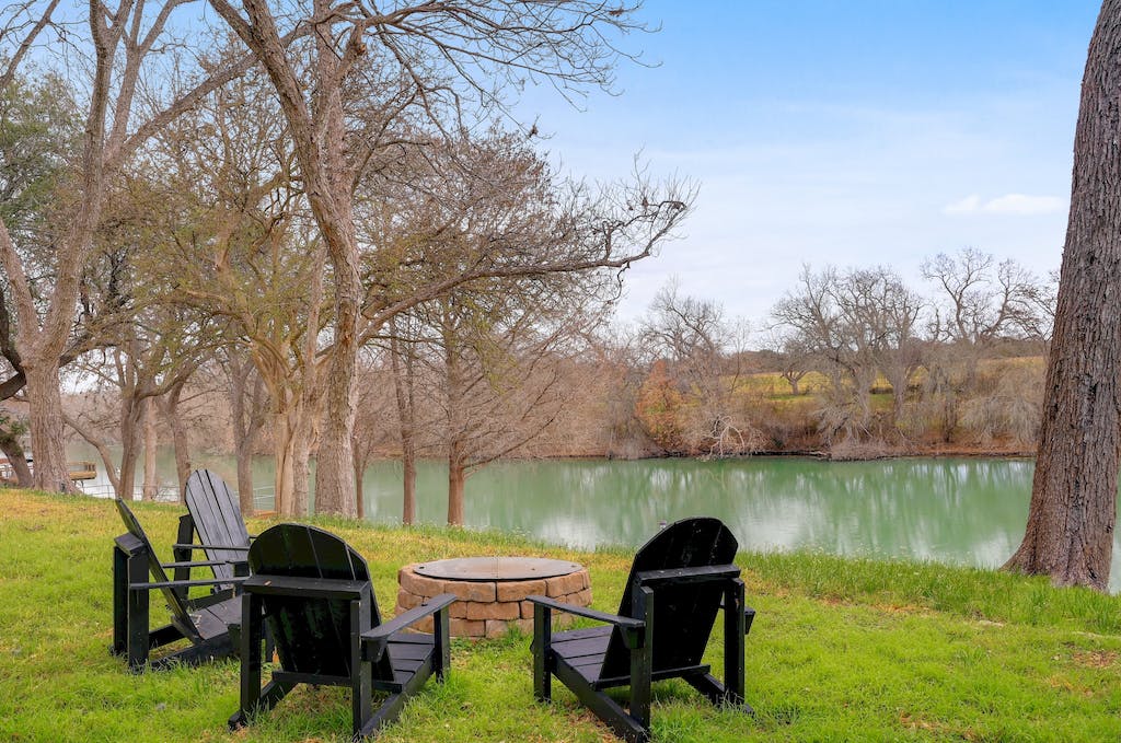 A serene outdoor setting features four black Adirondack chairs arranged around a stone fire pit. The area overlooks a calm body of water, framed by trees with bare branches, providing a peaceful spot for relaxation.