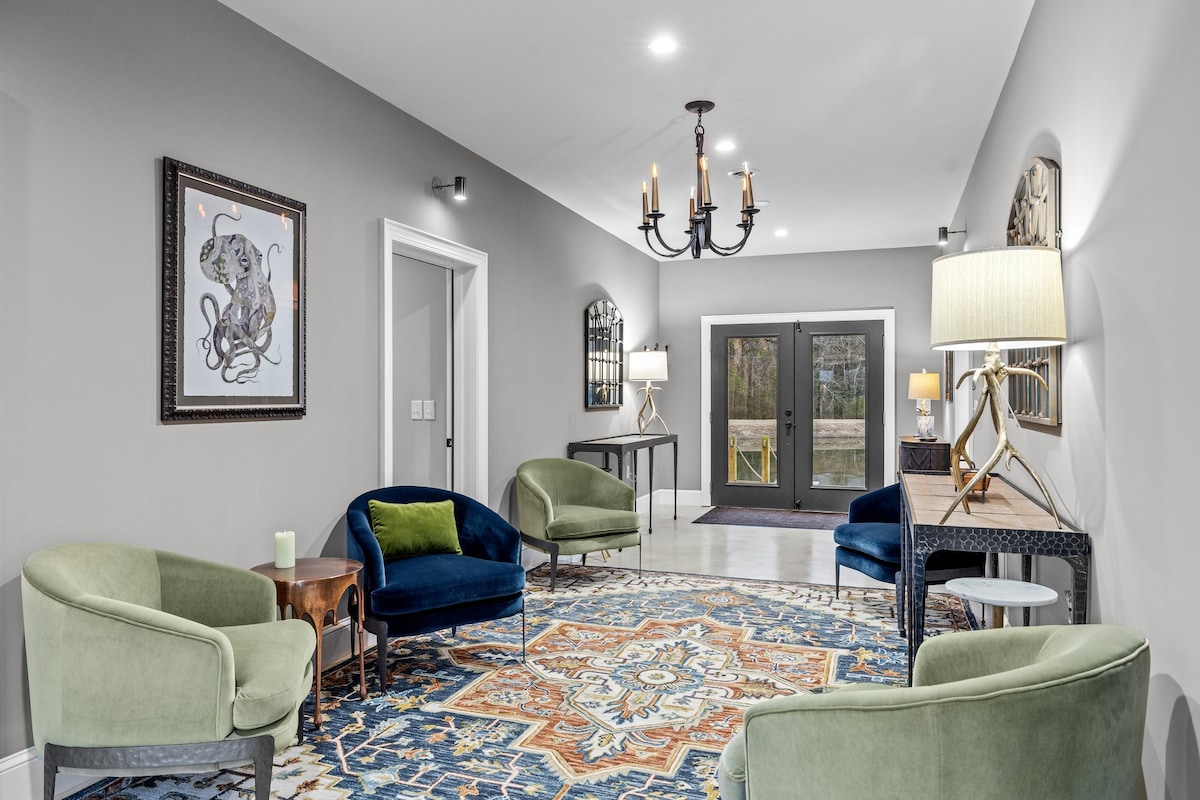 A spacious entryway is presented, featuring a colorful area rug at the center. Comfortable chairs in soft green offer seating, with a side table and stylish lighting enhancing the decor. Double doors lead to the outdoor area, while artwork adorns the walls.