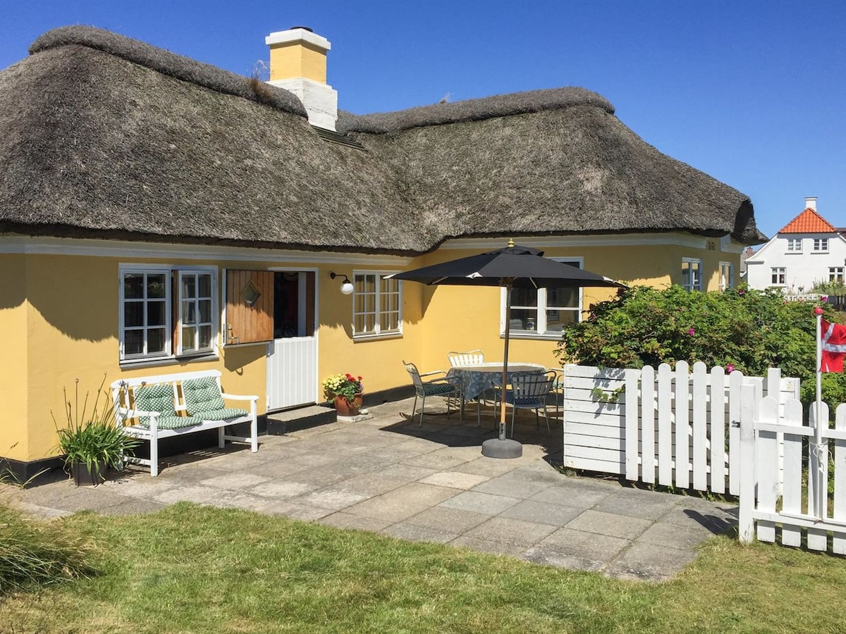 A charming single-family house features a thatched roof and yellow exterior. A spacious terrace includes a dining table and umbrella, surrounded by greenery. A white picket fence encloses the property, and comfortable seating is visible, enhancing the outdoor living space.