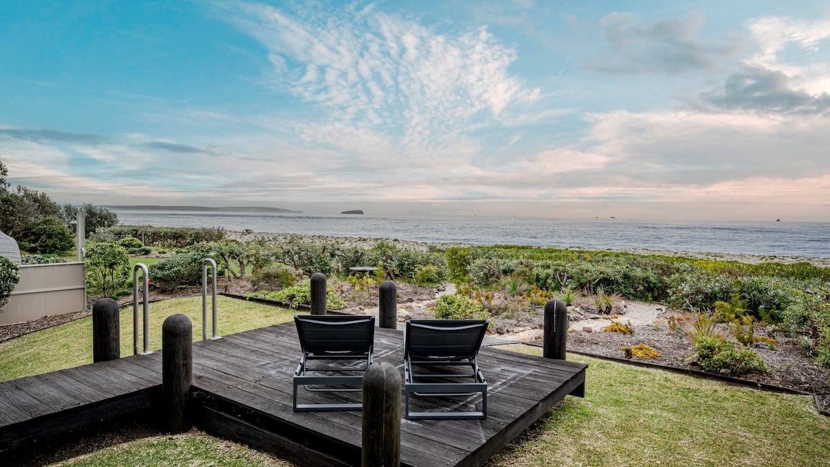 A wooden deck with two lounge chairs overlooks the beachfront and the ocean. Lush vegetation frames the area, while the calm waters extend to the horizon under a sky with soft clouds. The setting evokes a serene outdoor relaxation space.