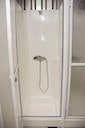 A modern shower is enclosed with clear glass doors. The shower features a chrome showerhead mounted on the wall, with a handheld option suspended nearby. The sleek, white tile flooring complements the minimalist design of the shower space.