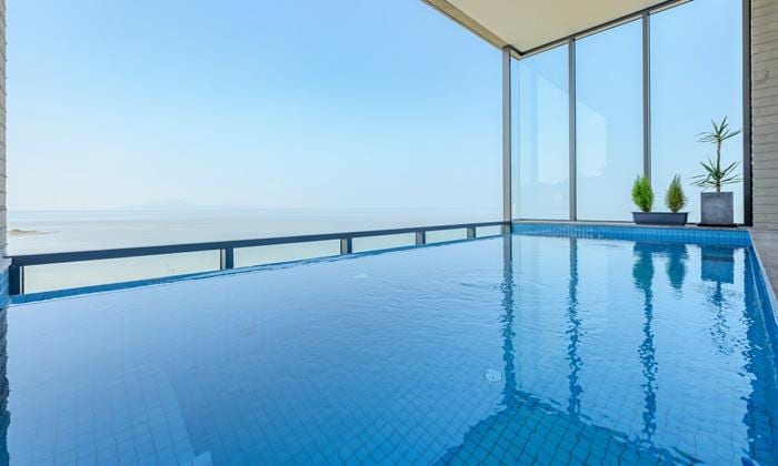 An infinity pool with tiled edges overlooks a vast ocean view, reflecting clear blue skies. Floor-to-ceiling windows provide unobstructed vistas, while potted greenery is positioned in the background, enhancing the serene atmosphere.