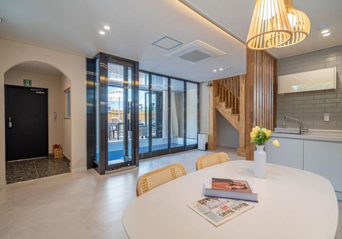 An open-concept living area features a round dining table with rattan chairs. Natural light floods in through large glass doors leading to an outdoor space. A modern kitchen is visible, along with a staircase leading to the upper level.