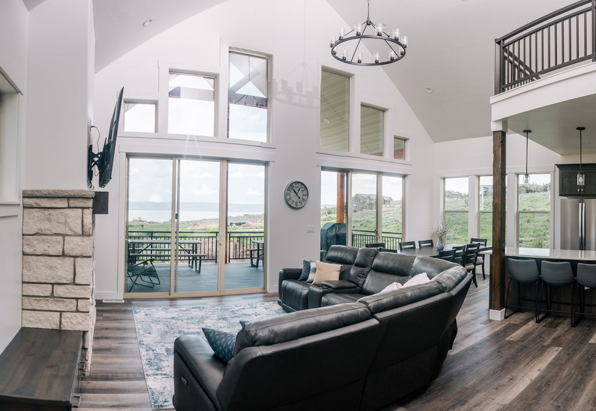 A spacious living room is highlighted by large windows that provide a view of the lake. A leather sectional faces an entertainment area with a wall-mounted television above a cozy fireplace. The open layout leads to a dining area and modern kitchen, enhancing the communal feel.