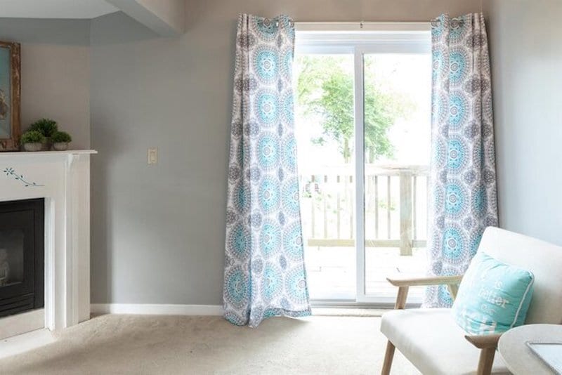 A cozy corner is created with a light chair positioned near a fireplace. Soft blue and patterned curtains frame a sliding door that leads to a deck, allowing natural light to brighten the neutral-colored walls.
