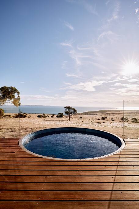 A heated plunge pool sits atop a wooden deck, providing serene views of the surrounding landscape. Soft sunlight shines over the horizon, enhancing the tranquil setting. The expansive vista includes rolling hills and coastal views, inviting relaxation and connection with nature.