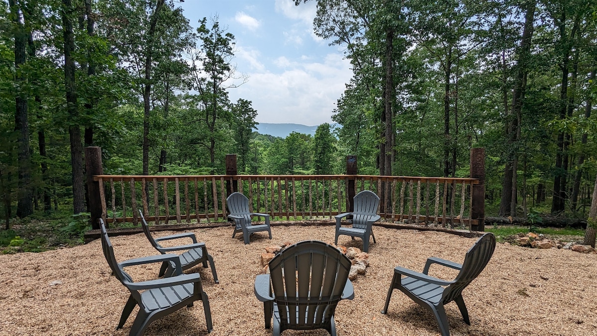 A gravel area is furnished with several gray lounge chairs arranged around a central space. A wooden railing provides a view of the surrounding forest and mountains, creating a serene outdoor setting.