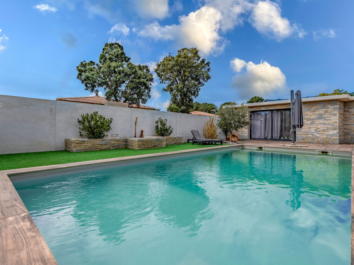 The swimming pool is surrounded by a well-maintained garden, featuring a mix of greenery and stone accents. A spacious deck area with lounge chairs is visible, alongside a shaded seating area. The pool's water appears inviting under a clear blue sky, with trees framing the background.