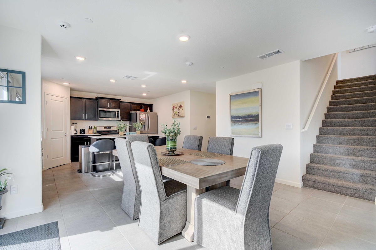 A modern dining area with a large wooden table surrounded by six upholstered chairs is visible. The space is filled with natural light, and an open layout connects it to a well-equipped kitchen. A staircase is featured in the background, leading to the upper floors.