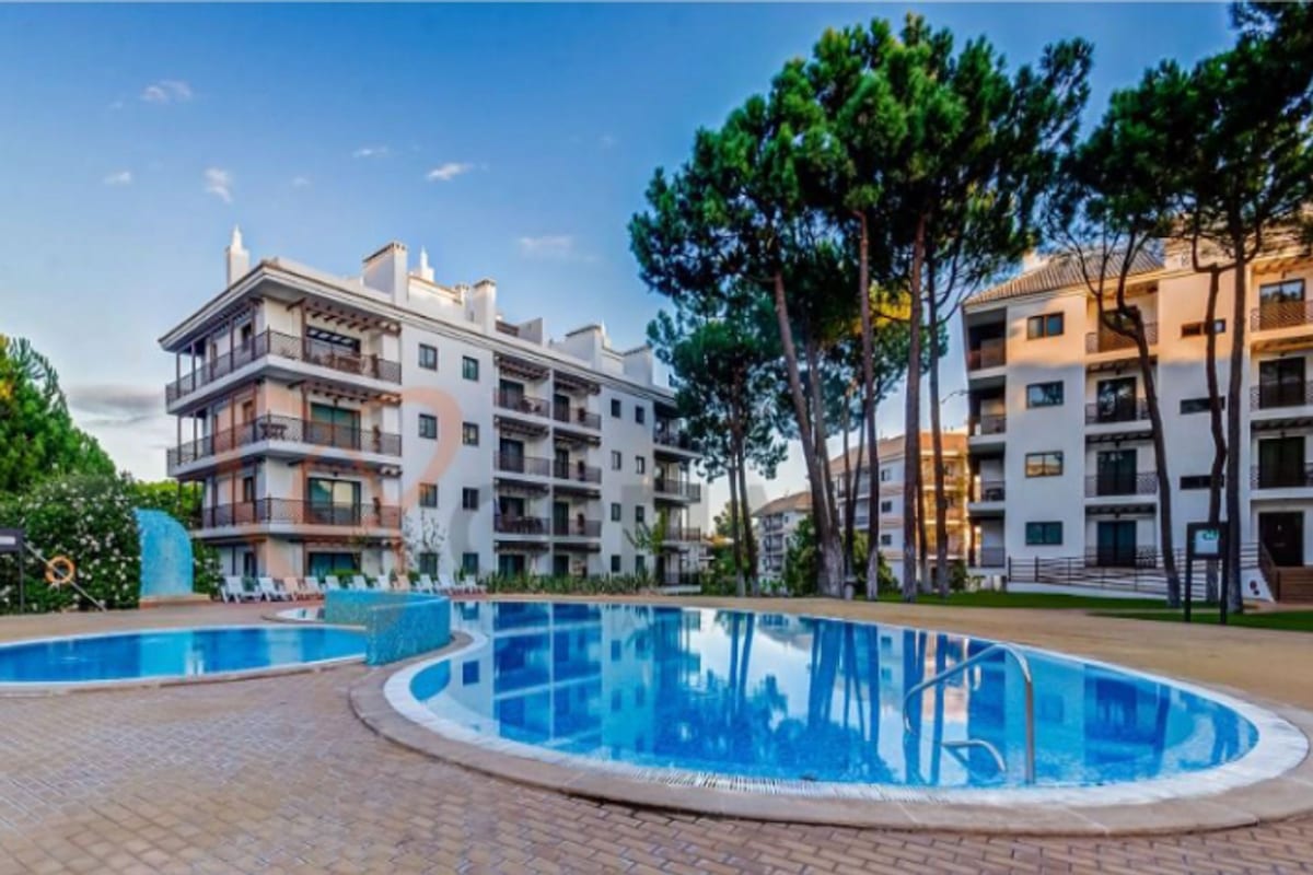 An outdoor swimming pool is framed by manicured trees and spacious buildings. The pool’s clear blue water reflects the sky, creating a serene environment. Surrounding areas feature neatly arranged patio spaces for lounging or relaxation.
