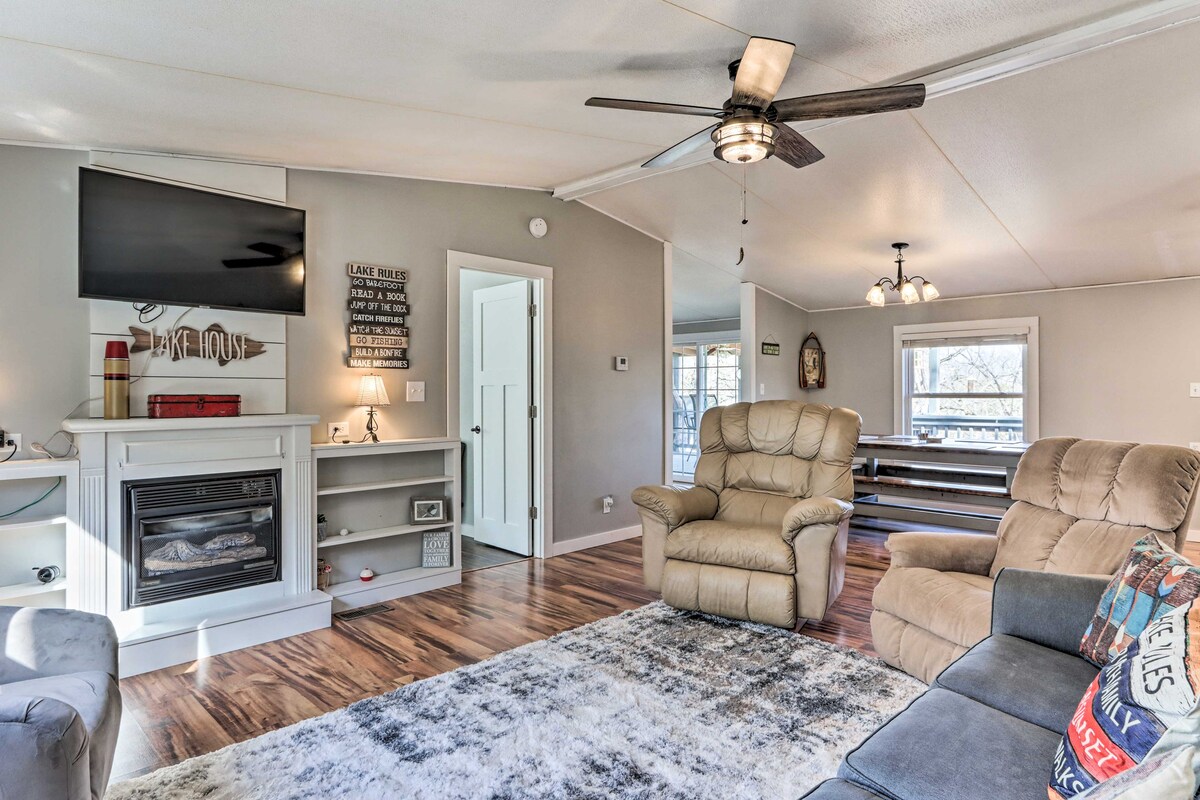 A welcoming living area features comfortable seating with tan recliner chairs and a gray sectional sofa. A flat-screen TV is mounted above a decorative fireplace, surrounded by wooden shelves. Natural light fills the space through nearby windows, enhancing the cozy ambiance.