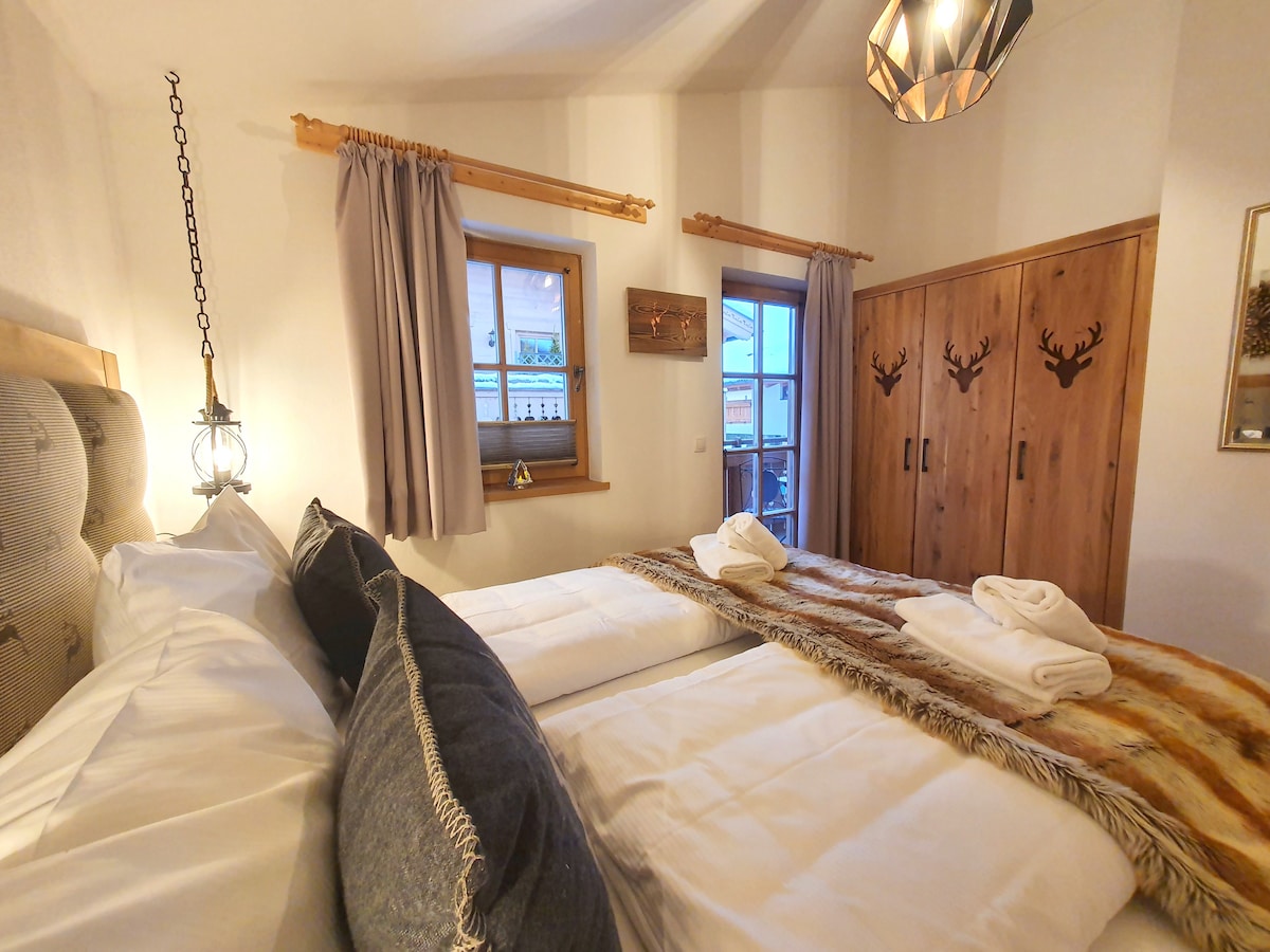 A serene bedroom features two spacious beds with crisp white linens and decorative throws. Soft natural light enters through two windows adorned with light curtains. Wooden wardrobes with decorative antlers add a rustic touch, complemented by a stylish pendant light overhead.