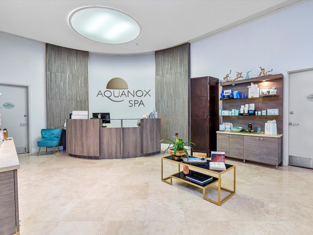 The spa reception area features a sleek, modern design with neutral tones. A circular overhead light brightens the space, while a organized display cabinet holds various spa products. A welcoming blue chair is positioned near the front desk, creating an inviting atmosphere.