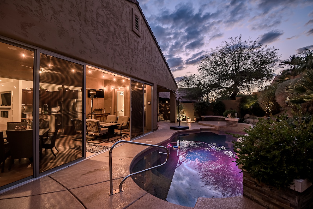 The exterior of the home is showcased at dusk, with oversized sliding glass doors reflecting warm indoor lighting. A private pool is visible, featuring a gentle glow from underwater lights, surrounded by lush landscaping and a cozy outdoor seating area.