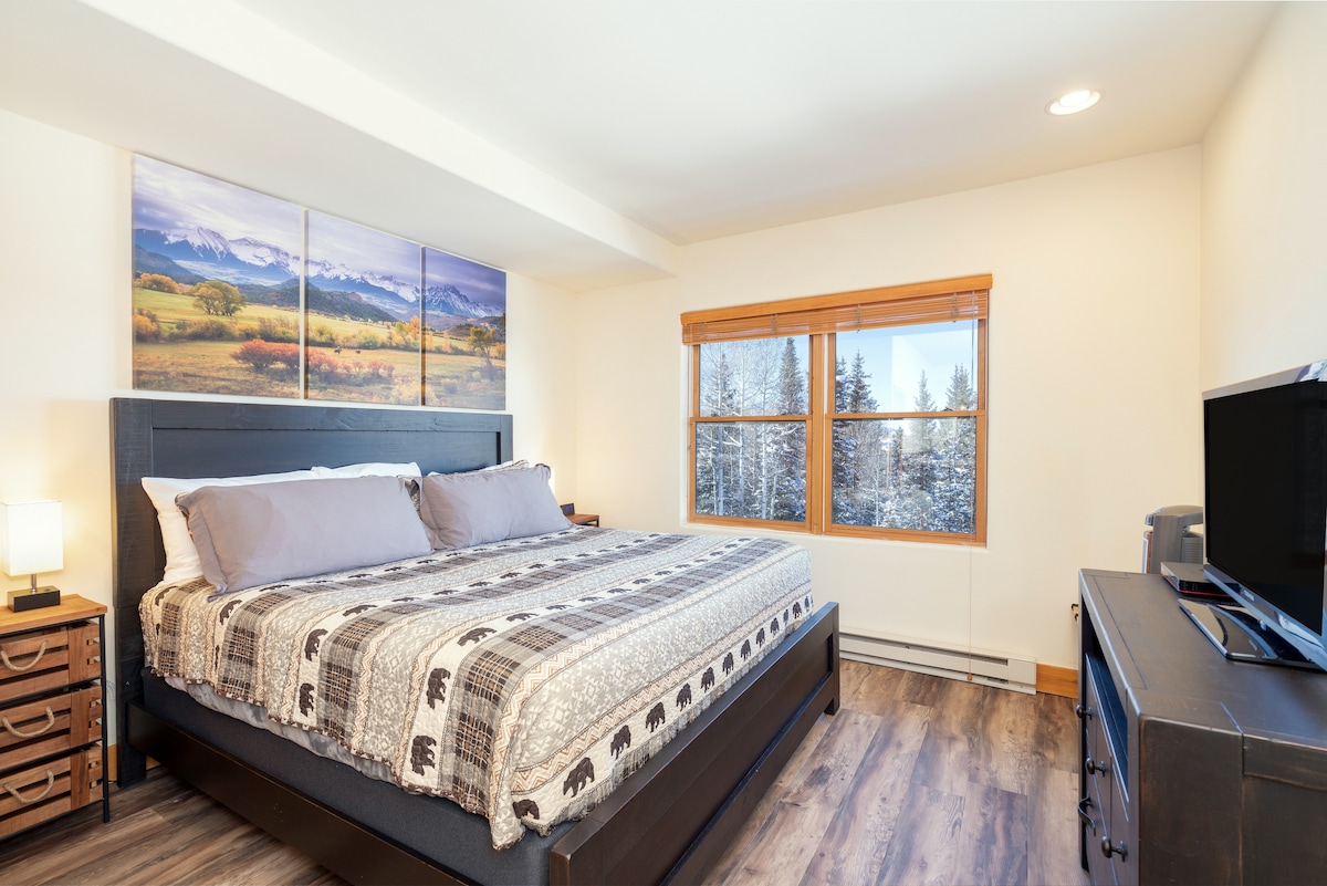 A spacious hotel-style room features a king bed adorned with a patterned blanket. Large windows provide natural light and a view of the snowy landscape outside. A flat-screen TV and small furniture are positioned near the bed for convenience.