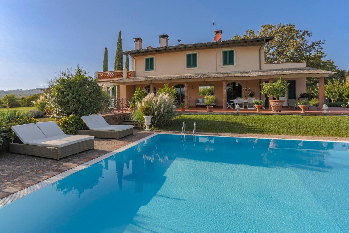 The villa is displayed with a serene swimming pool in the foreground, reflecting the clear sky. Comfortable sunbeds are positioned beside the pool, while the exterior of the two-story villa is framed by lush greenery and tall cypress trees.