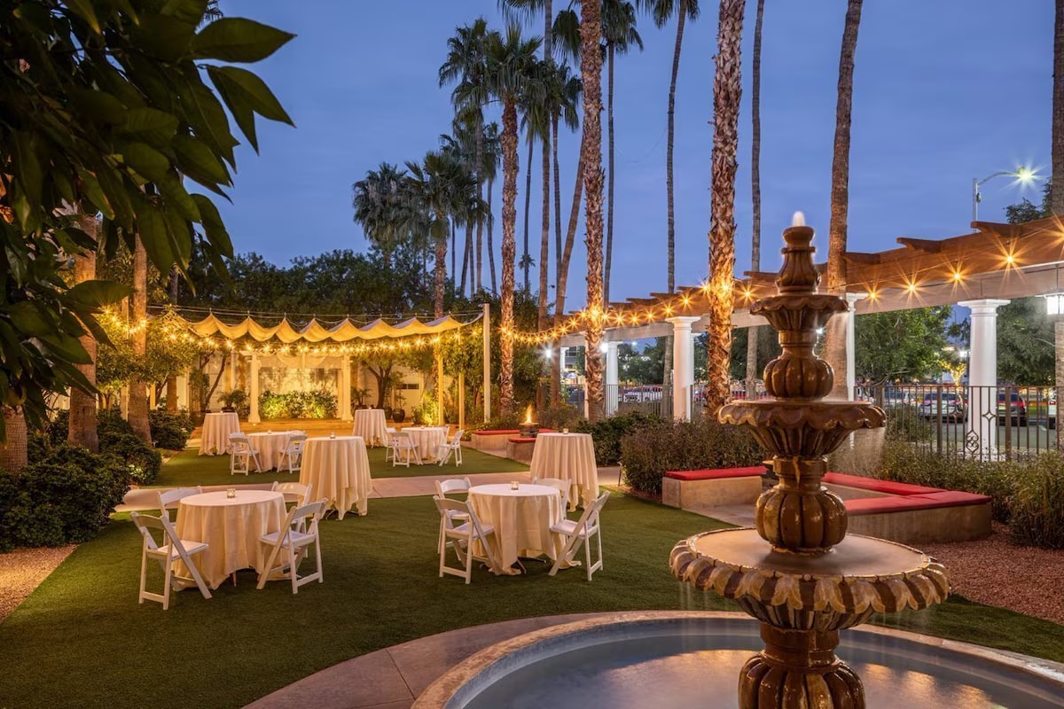 A serene outdoor space is highlighted by a decorative fountain in the foreground. Soft lighting drapes over tables covered with white tablecloths, set amidst lush greenery and palm trees. The setting creates a tranquil ambiance for gatherings or relaxation in the evening.