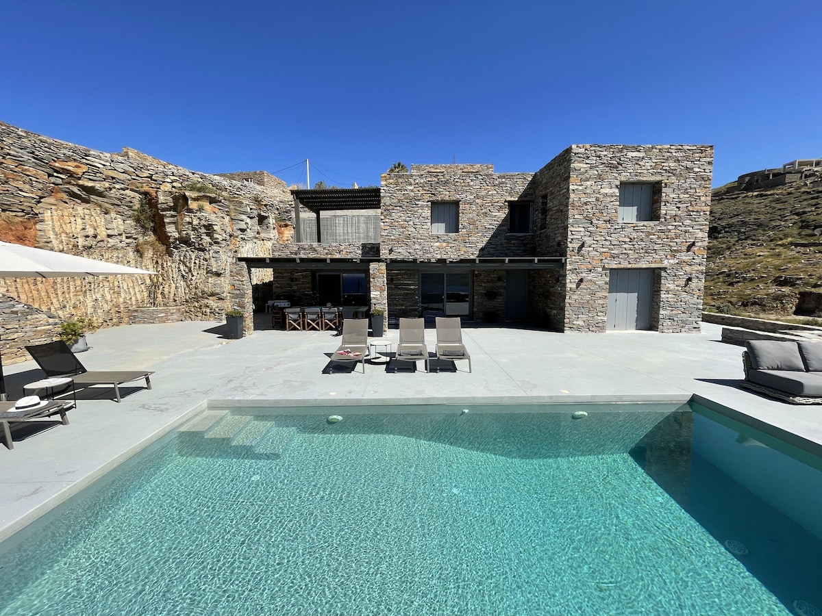 The villa's exterior is highlighted by a private swimming pool surrounded by loungers. Stone-clad walls complement the modern design, and wide glass doors open to an outdoor terrace area, providing access to the pool and dining space under shaded canopies.
