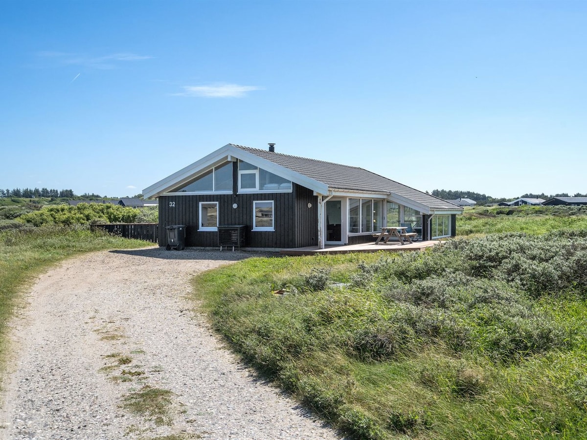 The single-family house is set in a natural landscape with surrounding greenery. The structure features a sloped roof and large windows, allowing ample sunlight. A gravel pathway leads to the entry, and a terrace is visible outside, suitable for outdoor seating.