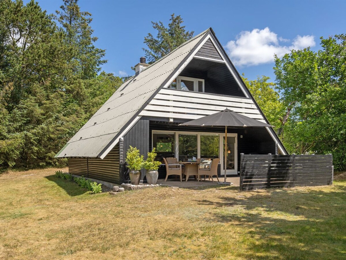 A single-family house with a distinct A-frame design is surrounded by natural landscaping. The exterior is a combination of black and white, featuring a spacious terrace with outdoor seating and an umbrella. Lush greenery and trees provide a serene backdrop.