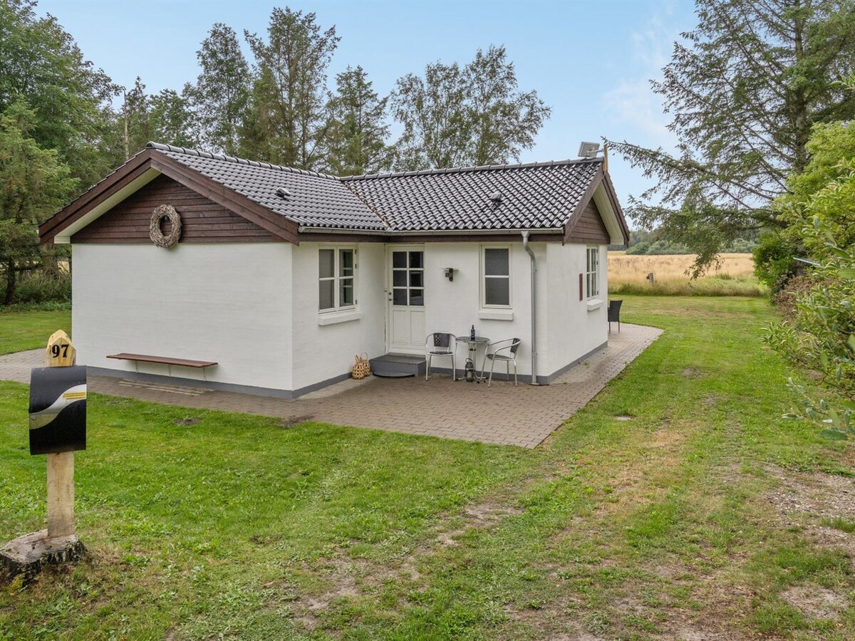 A single-family house is set within a spacious, well-kept garden. The exterior features a combination of white walls and wooden accents. A paved area is visible in front, with outdoor seating and a mailbox nearby, surrounded by greenery and open fields.