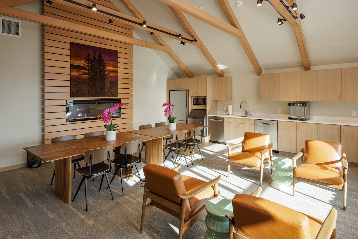 A spacious communal area is designed for comfort and relaxation, featuring a large wooden dining table surrounded by black chairs. Brown armchairs are positioned to create a cozy seating arrangement. A modern fireplace and kitchenette are visible, with a large artwork hanging on the wall.