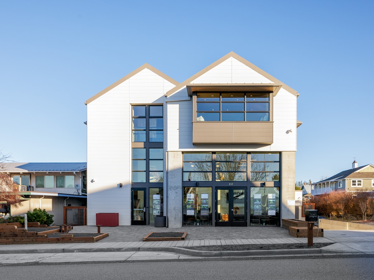 The exterior of the building features a modern design with large glass windows and a combination of white and gray paneling. The two-story structure includes a balcony on the upper level, framed by clean lines and a welcoming entrance. Surrounding landscaping is minimal and well-maintained.