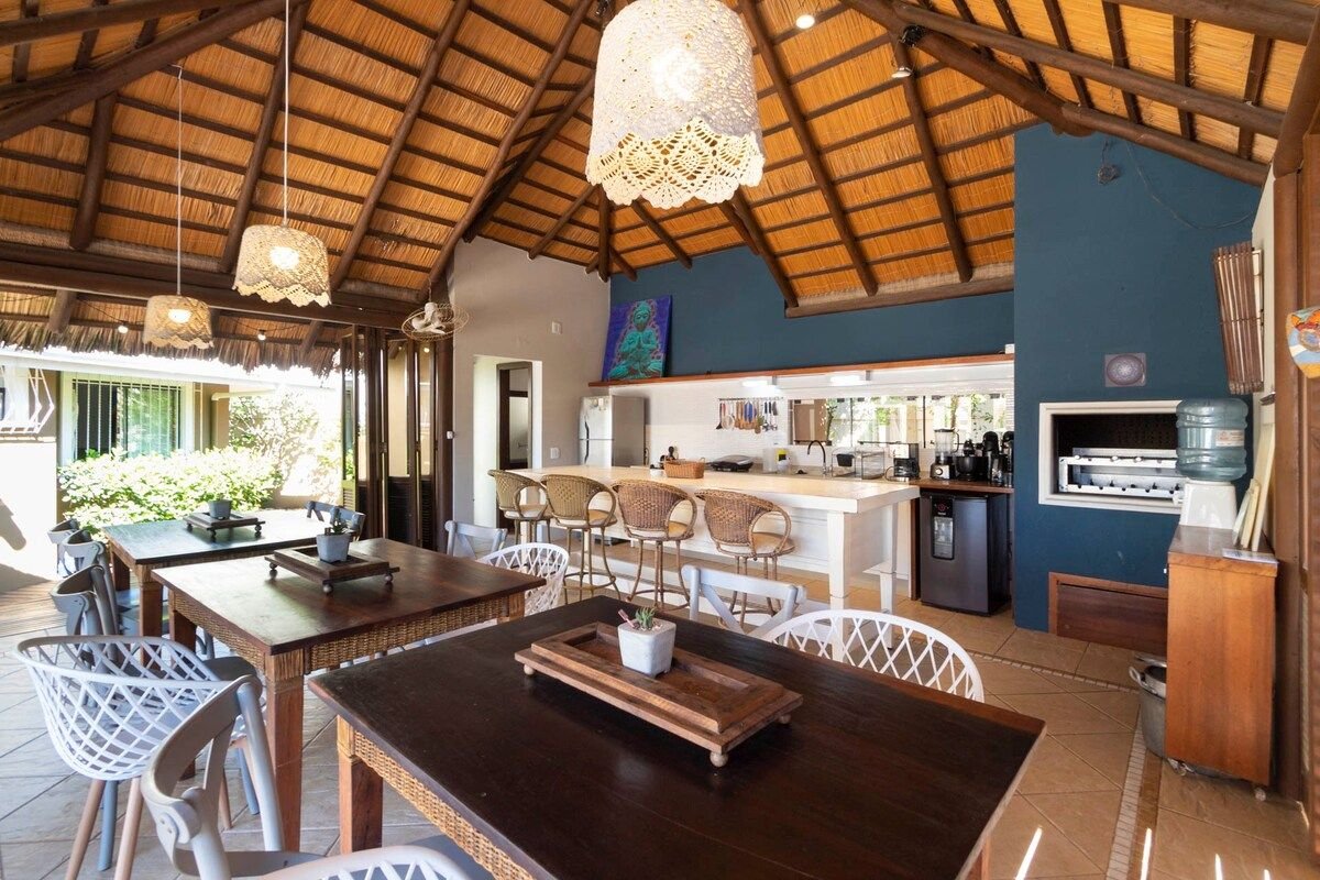 An open dining area features a high thatched ceiling, illuminated by woven pendant lights. Tables with white chairs are arranged around, and a bar area with stools integrates the space. A modern kitchen is visible in the background, enhancing the welcoming dining environment.