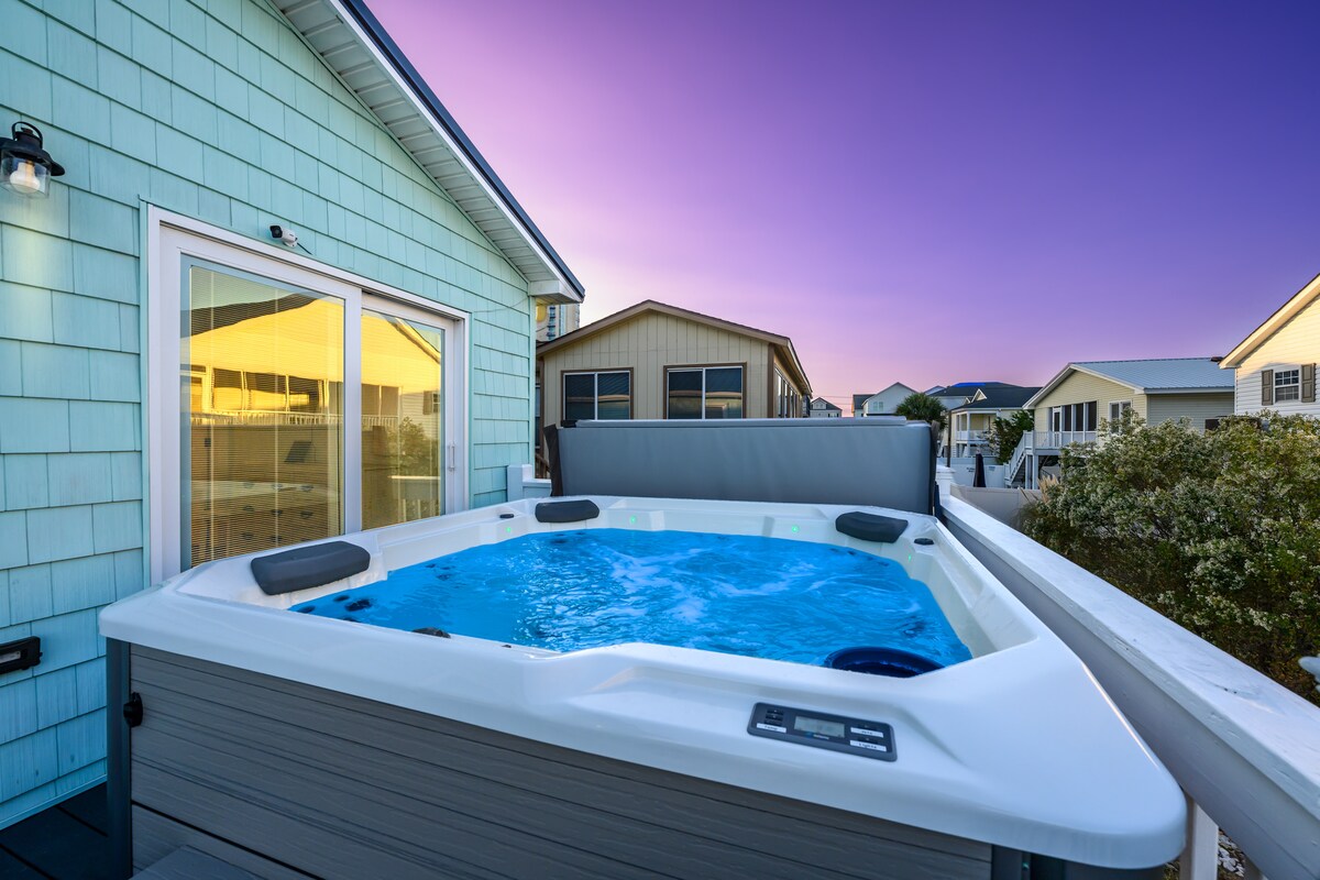 A newly installed hot tub is featured on the back porch, surrounded by a wooden railing. The water appears inviting under the twilight sky, reflecting shades of blue. Soft seating is positioned around the tub, enhancing the outdoor relaxation space.