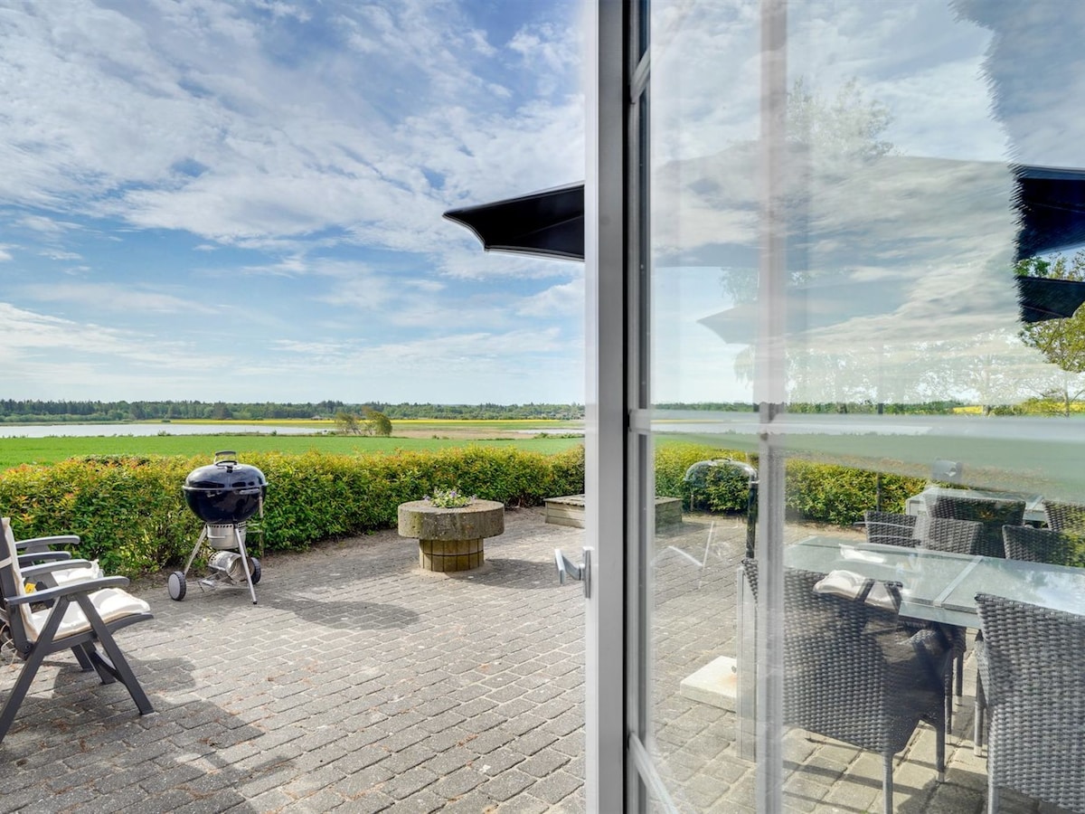 An outdoor terrace is showcased with a dining table and chairs, overlooking a scenic view of the water and lush greenery. A barbecue grill is positioned nearby, and the space is enhanced by the soft light reflecting off the window panes.
