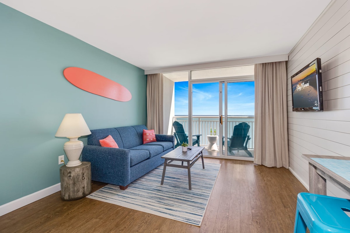 A bright living area features a blue sofa accented with coral-colored pillows. A coffee table sits on a striped area rug, complemented by a decorative surfboard on the wall. Large sliding doors offer access to a private balcony with ocean views and blue adirondack chairs.