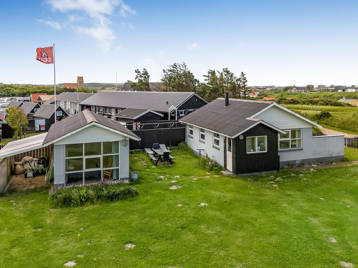 A spacious outdoor area is visible, featuring green grass and a mix of traditional and modern wooden structures. Flags fly gently in the breeze, and picnic tables are set under a shelter. Surrounding vegetation adds to the natural ambiance of the location.