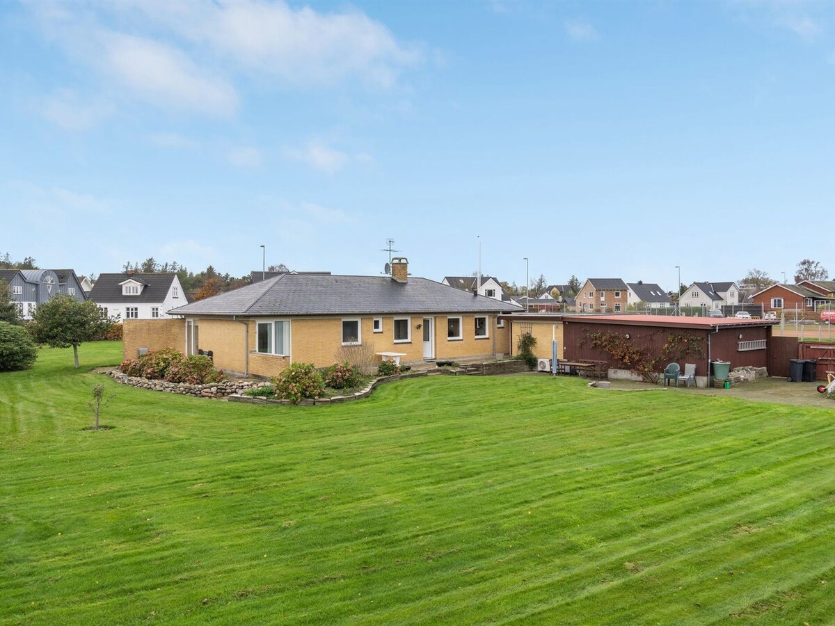 The exterior of the house displays a single-story structure with a light yellow facade, surrounded by a well-maintained green lawn. A fence encloses the property, creating a spacious yard. Nearby residential buildings provide a backdrop to this peaceful setting.