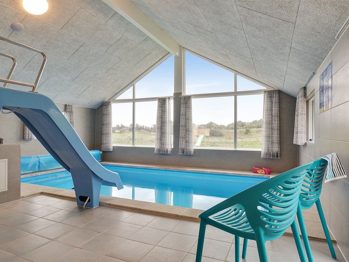 An indoor pool area features a blue water slide and a rectangular pool. Large windows bring in natural light and offer a view of the greenery outside. Two turquoise chairs are positioned along the poolside, enhancing the space's functionality.